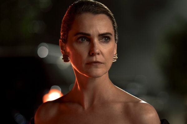 "Woman with slicked-back hair and elegant black dress stands outdoors at night, dramatic lighting highlighting her serious expression, blurred background with plants and car."
