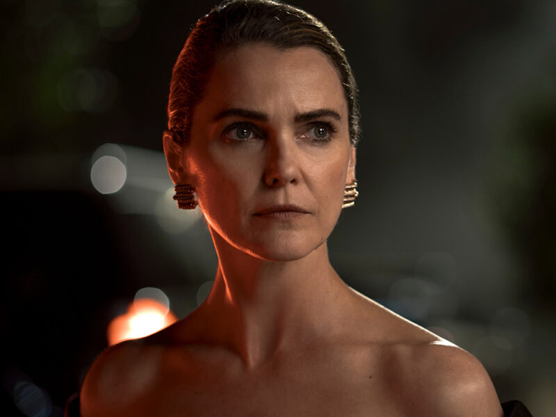 "Woman with slicked-back hair and elegant black dress stands outdoors at night, dramatic lighting highlighting her serious expression, blurred background with plants and car."